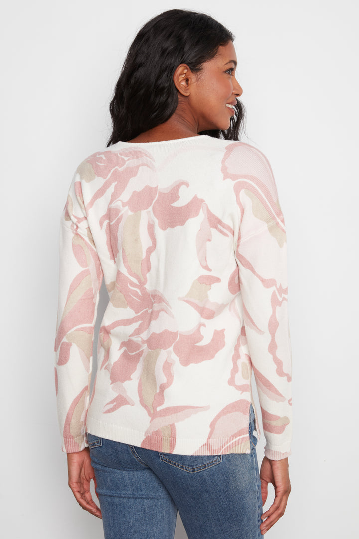 A woman shown from the back wearing a long-sleeve cream knit top with a pink and beige abstract floral pattern, paired with blue jeans, against a light gray studio background.