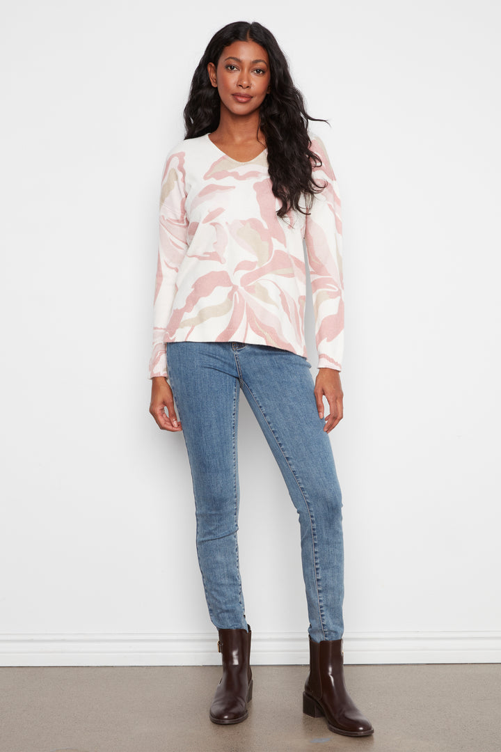 A woman modeling a long-sleeve V-neck top with a pink and white abstract floral pattern, paired with blue jeans and dark brown ankle boots, standing against a white wall.