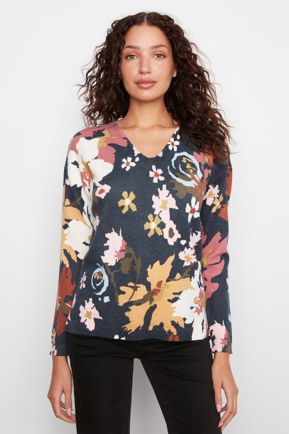 A woman with curly brown hair wearing a long-sleeve V-neck top with a multicolor floral and abstract print on a dark navy base, paired with black pants, against a light gray studio background.