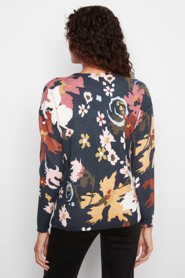 A woman viewed from behind wearing a long-sleeve knit top with a dark navy base and a large multicolor floral pattern in pink, orange, yellow, and white, paired with black pants against a light gray studio background.