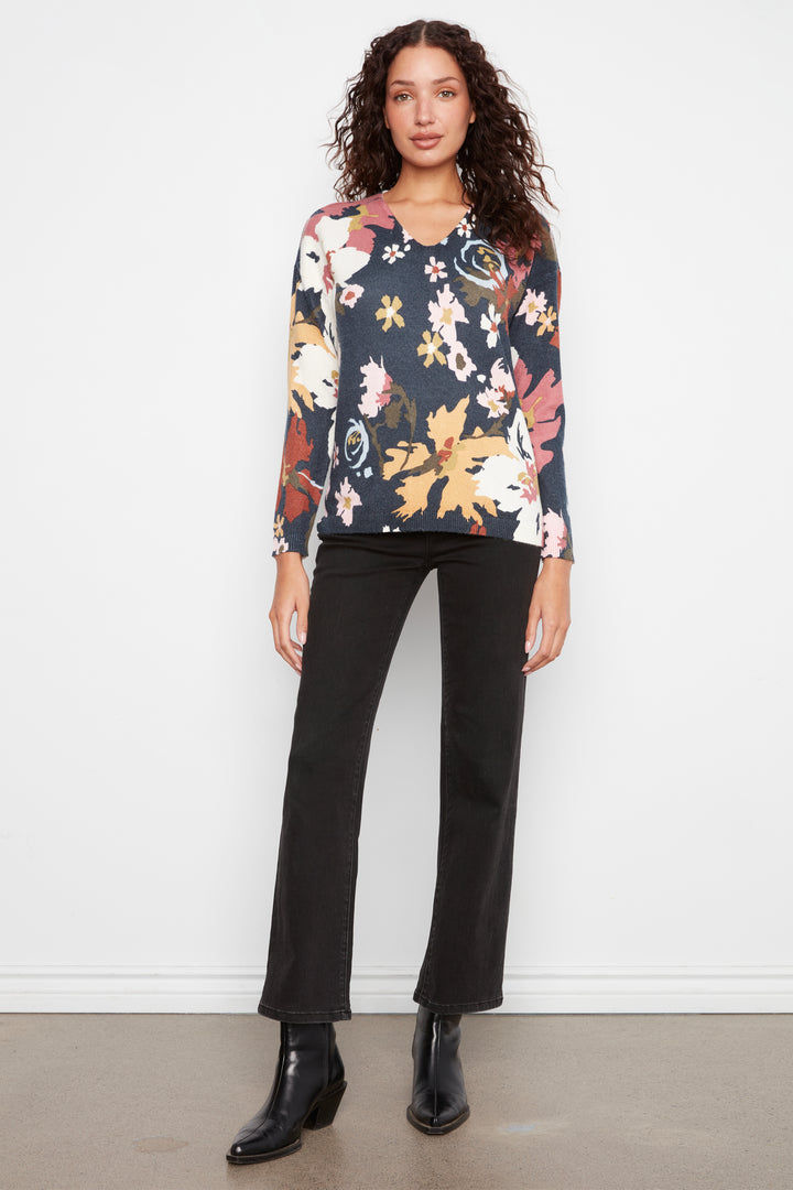 A woman modeling a long-sleeve floral blouse with a dark navy base and multicolor floral print, paired with black pants and black ankle boots, standing against a white wall.