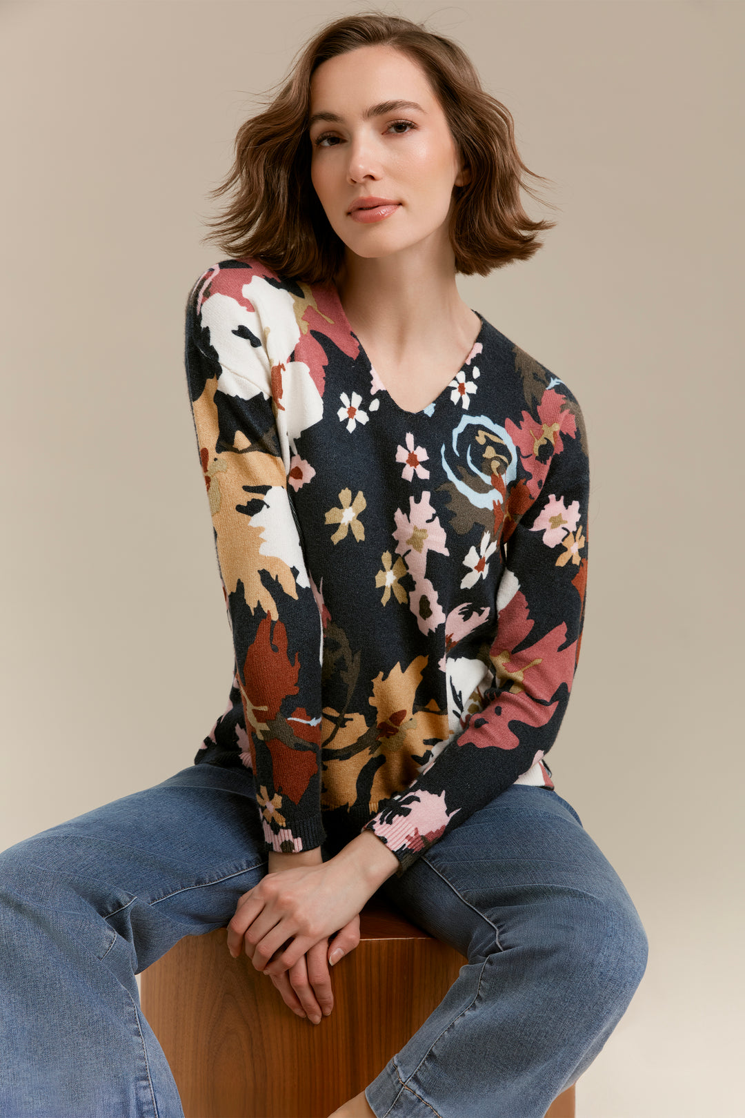 A woman with short brown hair sits on a wooden stool wearing a long-sleeve V-neck knit sweater with a multicolor floral pattern on a dark background, paired with blue jeans, against a solid beige studio backdrop.