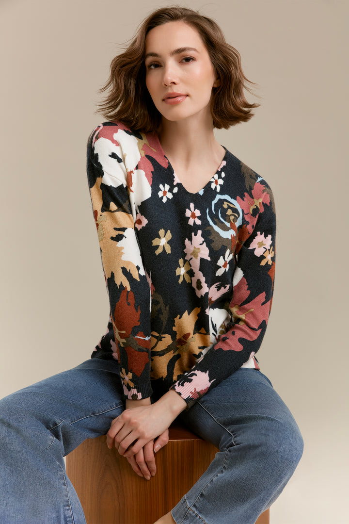 A woman with short brown hair sits on a wooden stool wearing a long-sleeve V-neck knit sweater with a multicolor floral pattern on a dark background, paired with blue jeans, against a solid beige studio backdrop.
