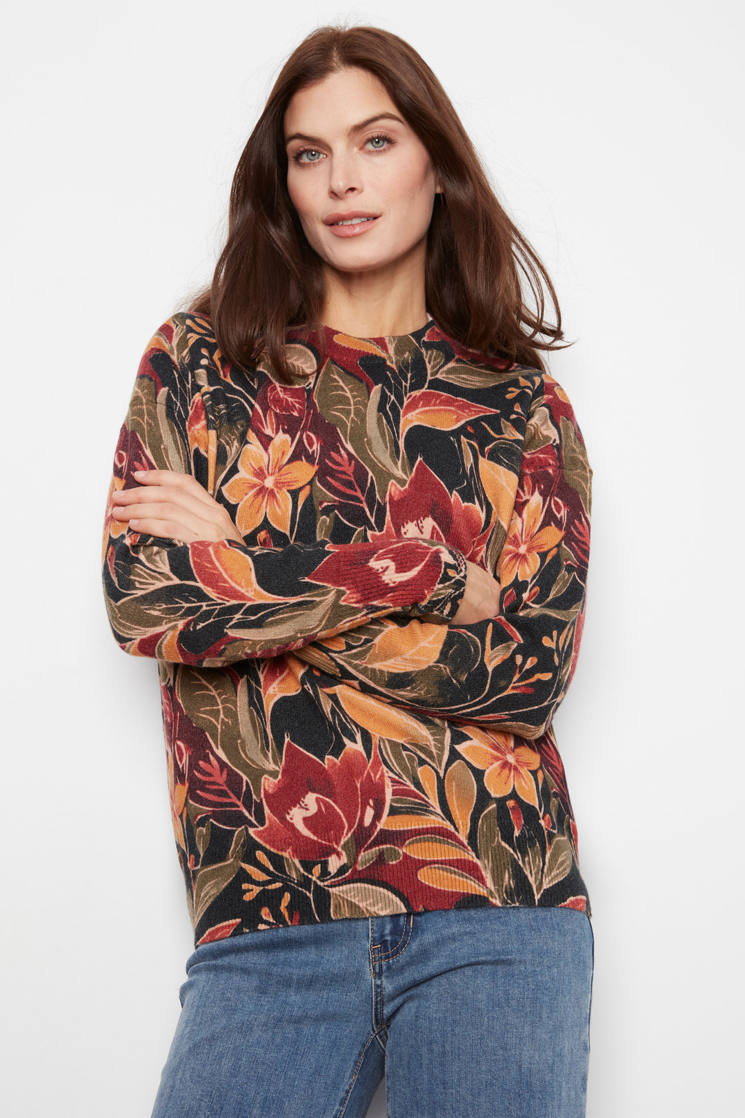A woman wearing a long-sleeve crewneck top with a dense tropical floral print in red, orange, and brown tones over a dark background, paired with blue jeans, against a plain white studio background.