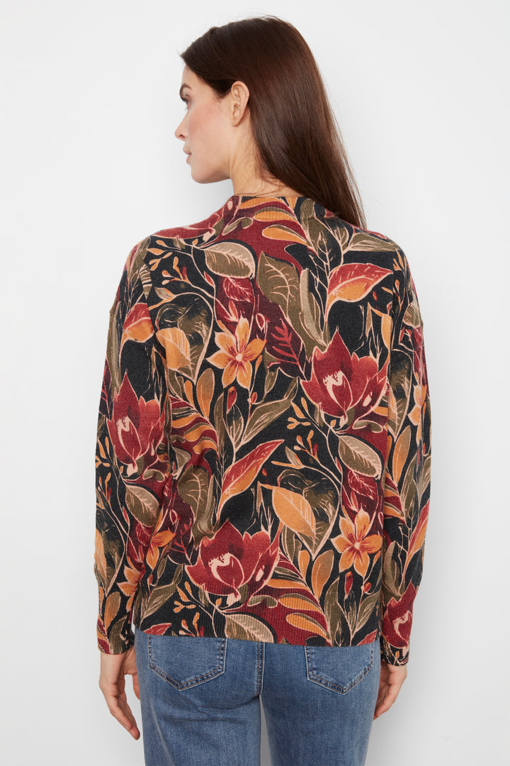 A woman shown from the back wearing a long-sleeve blouse with a dense floral and botanical print in red, orange, brown, and black, paired with blue jeans against a plain light gray studio background.