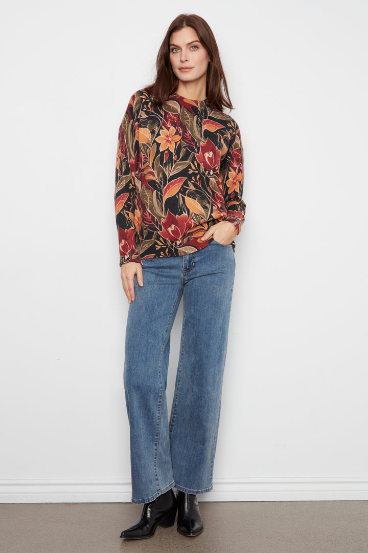 A woman stands against a plain white wall wearing a long-sleeve floral print top with blue wide-leg jeans and black ankle boots.
