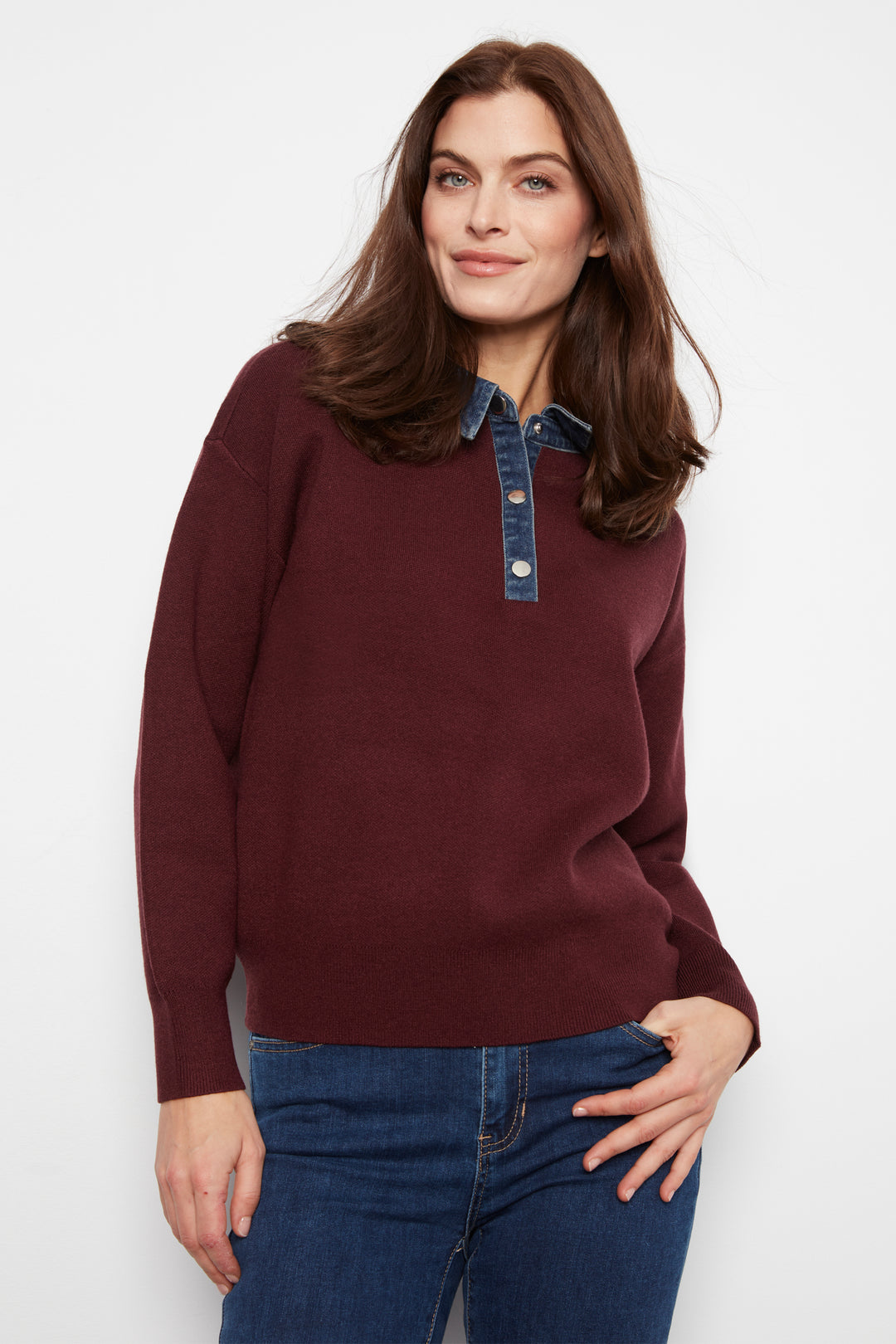 A woman wearing a long-sleeve knit polo-style sweater in deep burgundy with a contrasting blue denim collar and button placket, paired with blue jeans, against a plain white studio background.