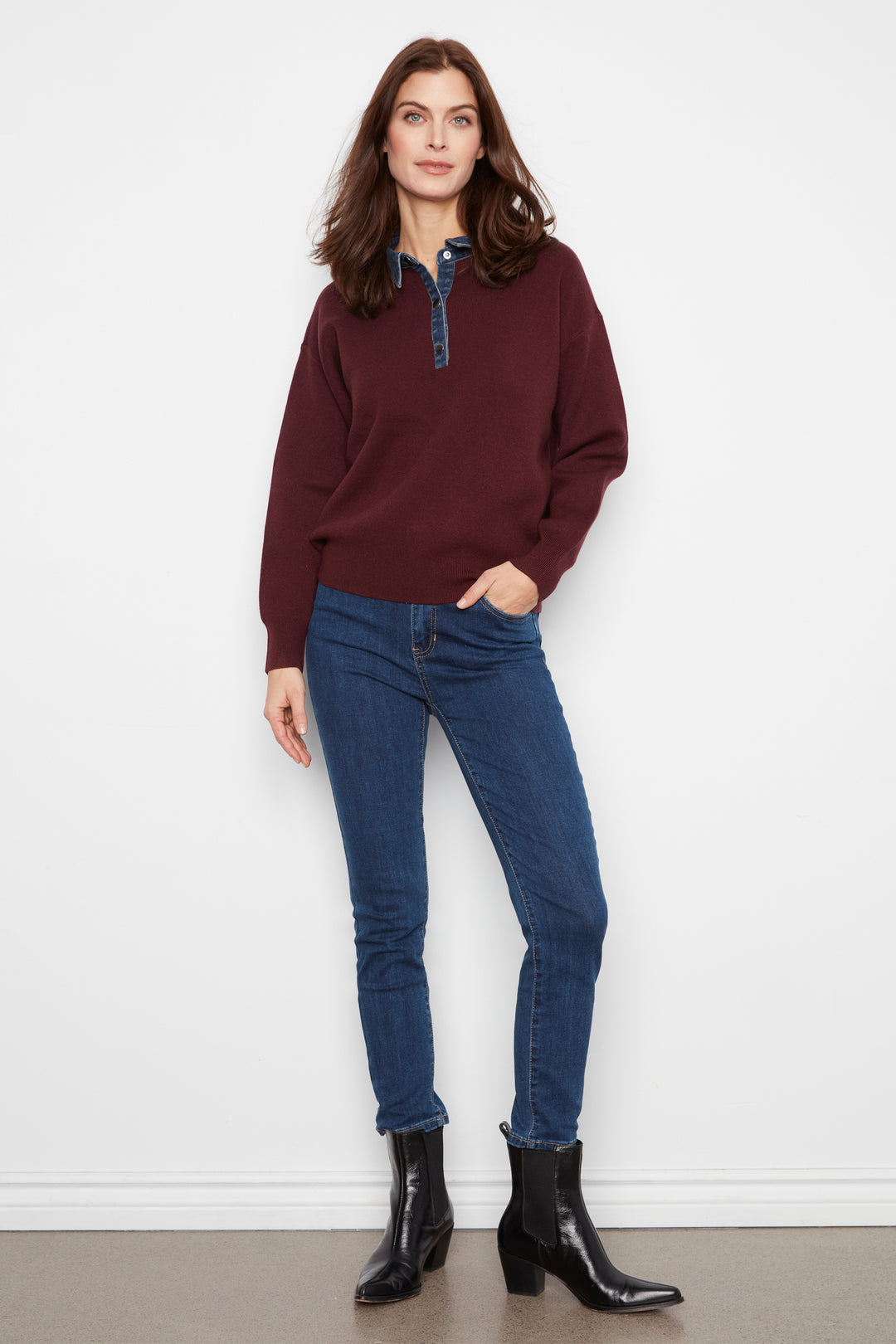 A woman modeling a burgundy long-sleeve quarter-placket sweater with a blue patterned collar detail, paired with dark blue skinny jeans and black ankle boots, standing against a white wall.