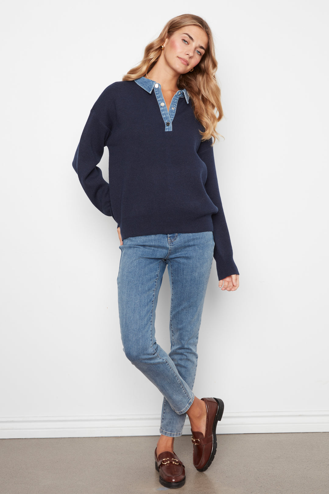 A woman modeling a navy blue long-sleeve knit polo sweater with a light blue denim collar detail, paired with light-wash blue jeans and brown leather loafers, standing against a white wall.