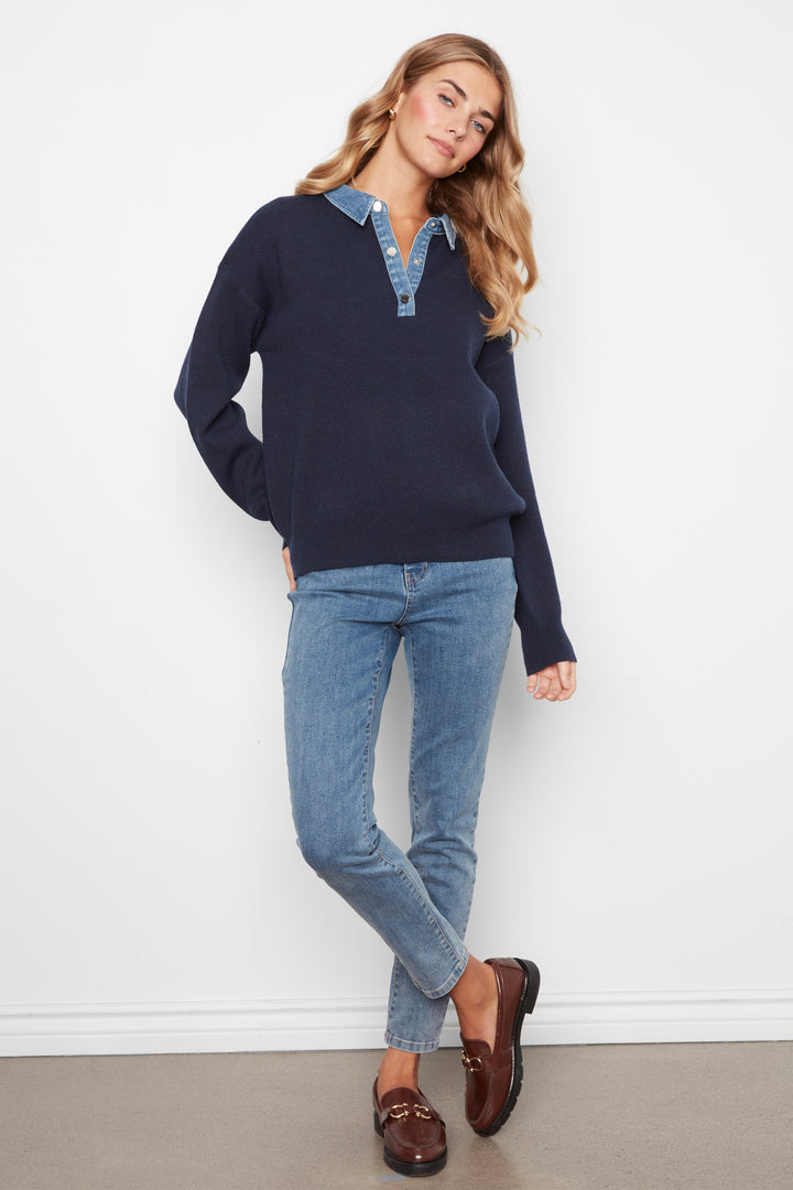 A woman modeling a navy blue long-sleeve knit polo sweater with a light blue denim collar detail, paired with light-wash blue jeans and brown leather loafers, standing against a white wall.