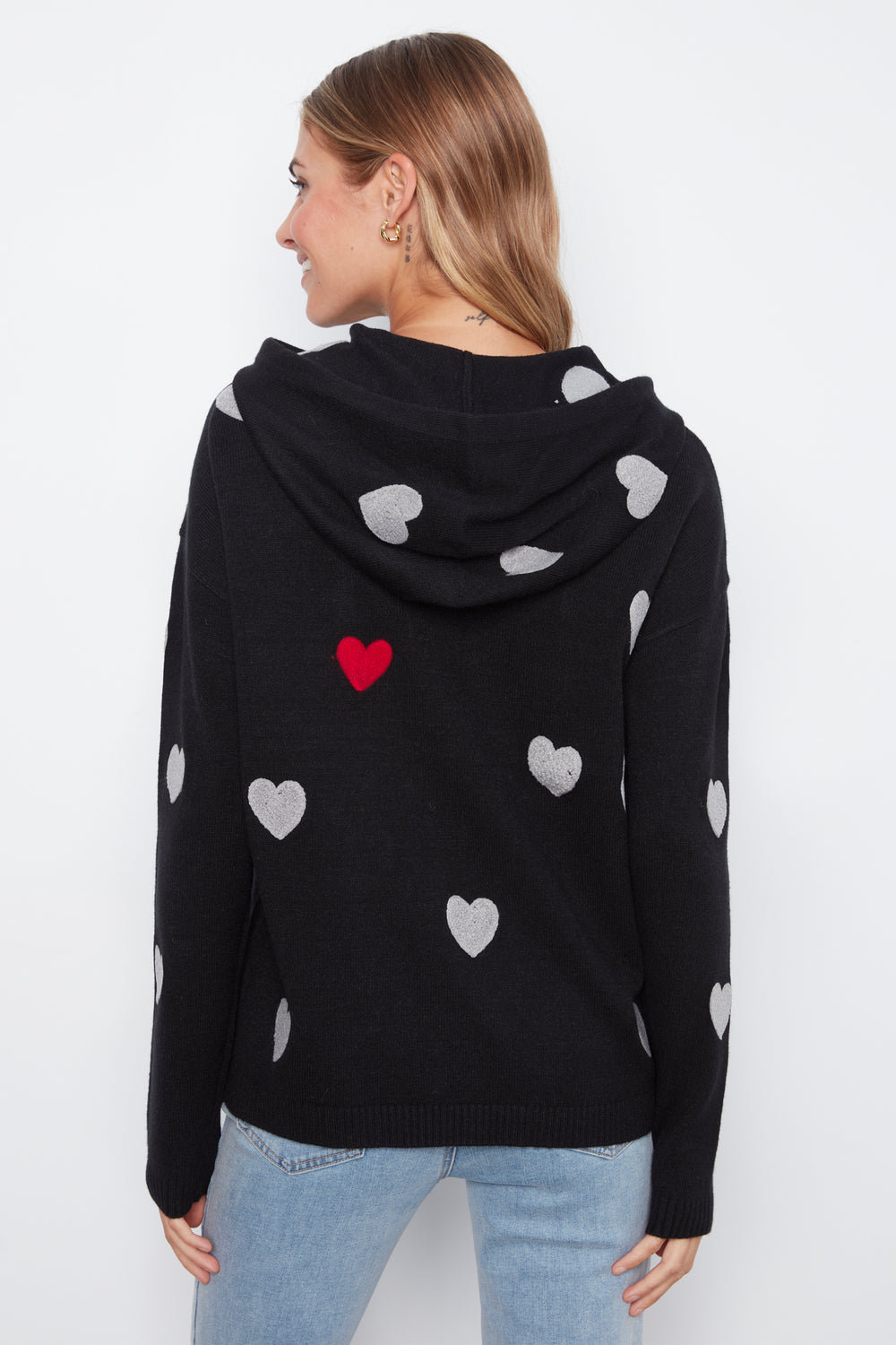 A woman shown from the back wearing a black knit hoodie with scattered white hearts and one red heart on the back, paired with light blue jeans against a plain white background.