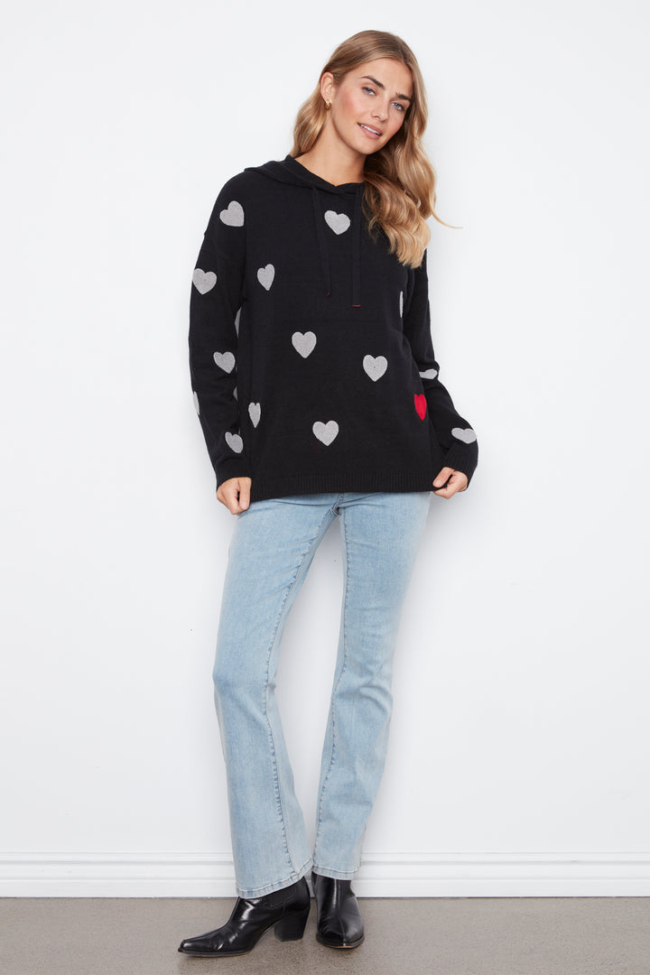 A woman modeling a black long-sleeve hoodie with a pattern of white hearts and a small red heart, paired with light-wash blue jeans and black ankle boots, standing against a white wall.