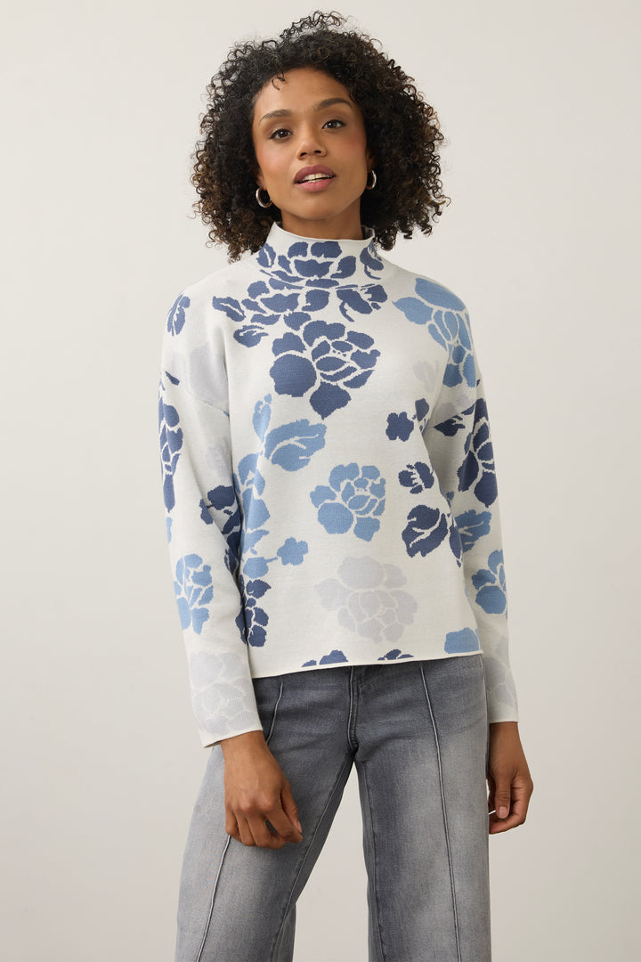 A woman modeling a white mock-neck knit sweater with a blue floral pattern, paired with gray jeans, against a plain light gray background.