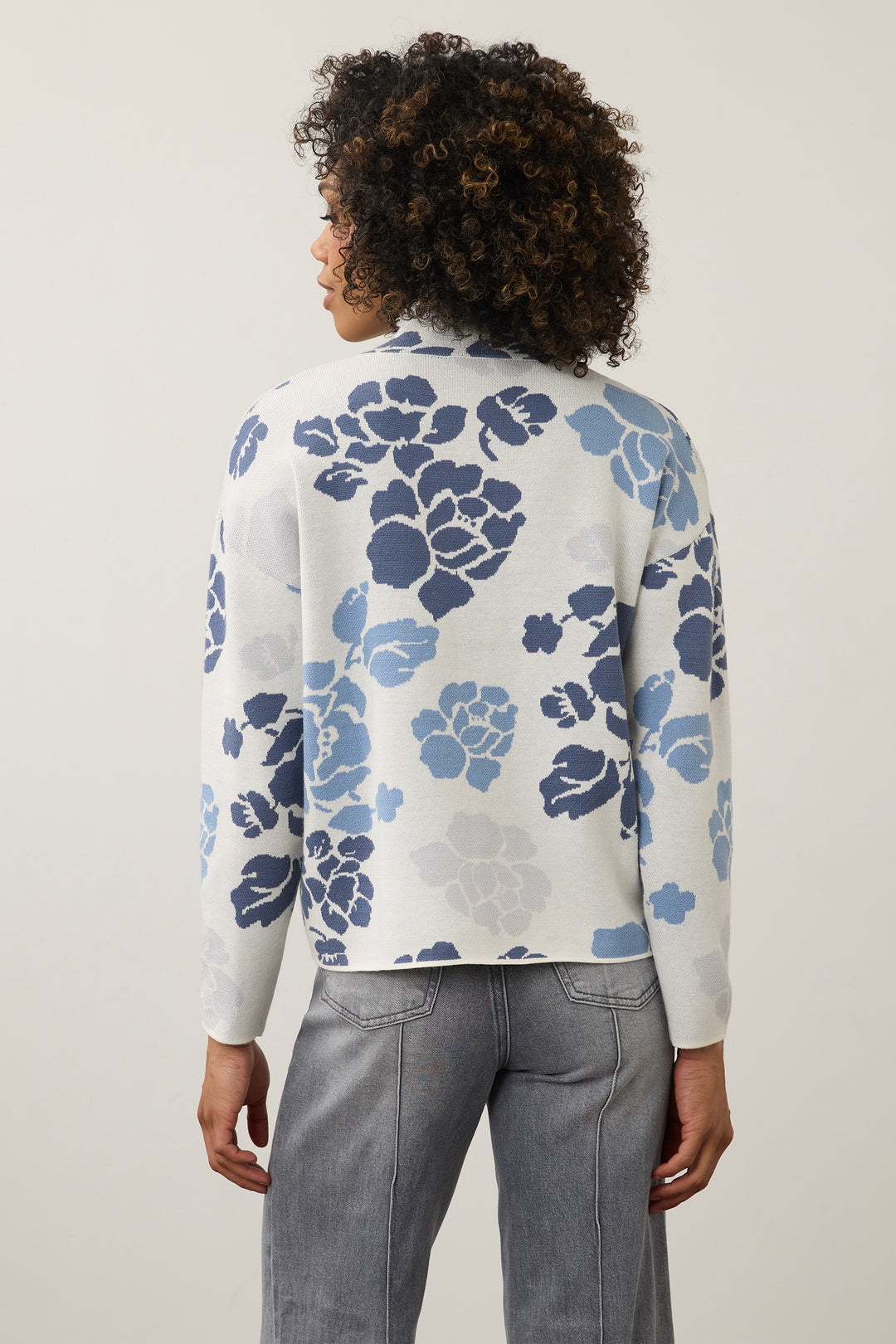 A woman modeling a white mock-neck knit sweater with a blue floral pattern, paired with wide-leg gray jeans and navy flats, standing against a plain off-white wall.