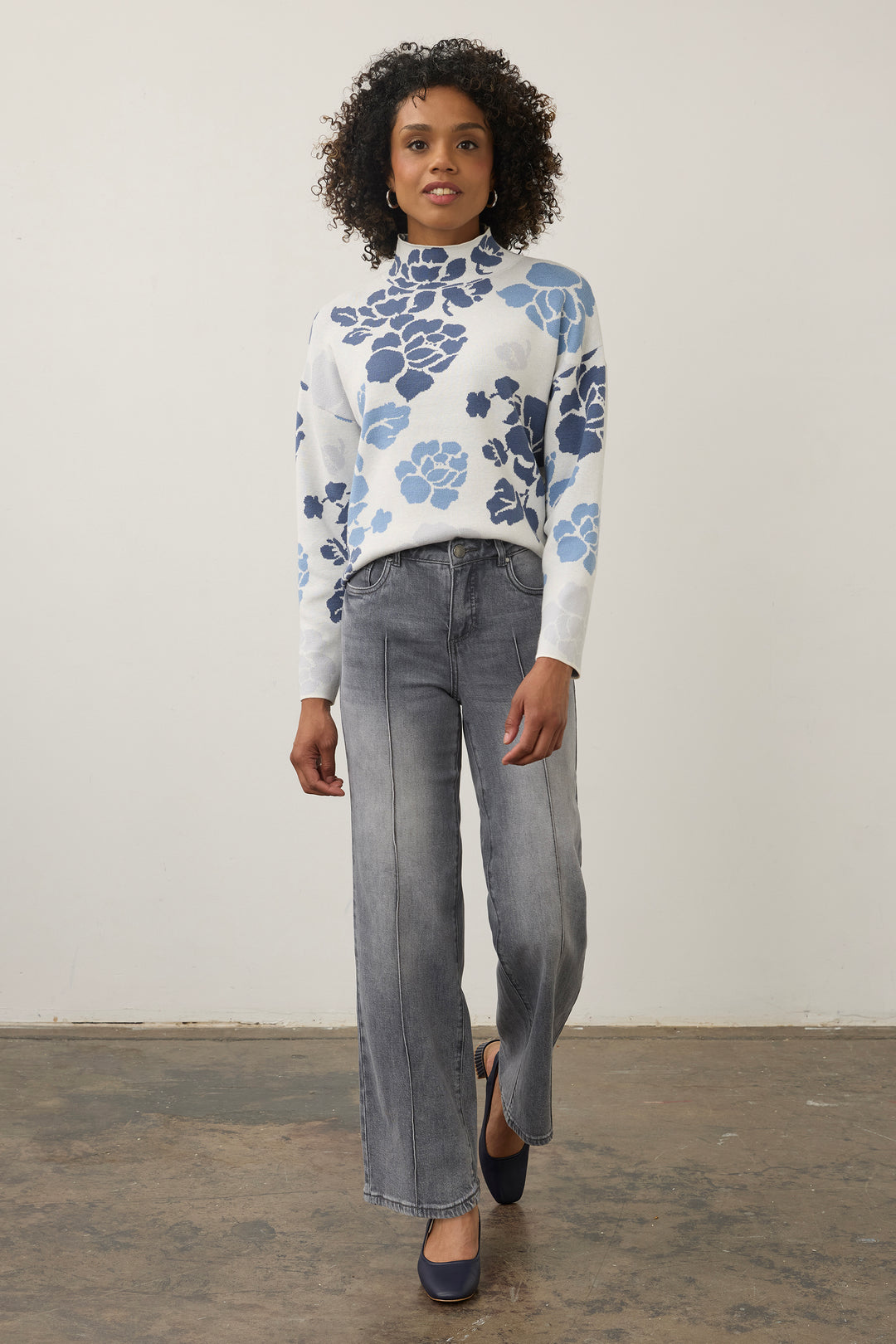 A woman modeling a white mock-neck knit sweater with a blue floral pattern, paired with wide-leg gray jeans and navy flats, standing against a plain off-white wall.
