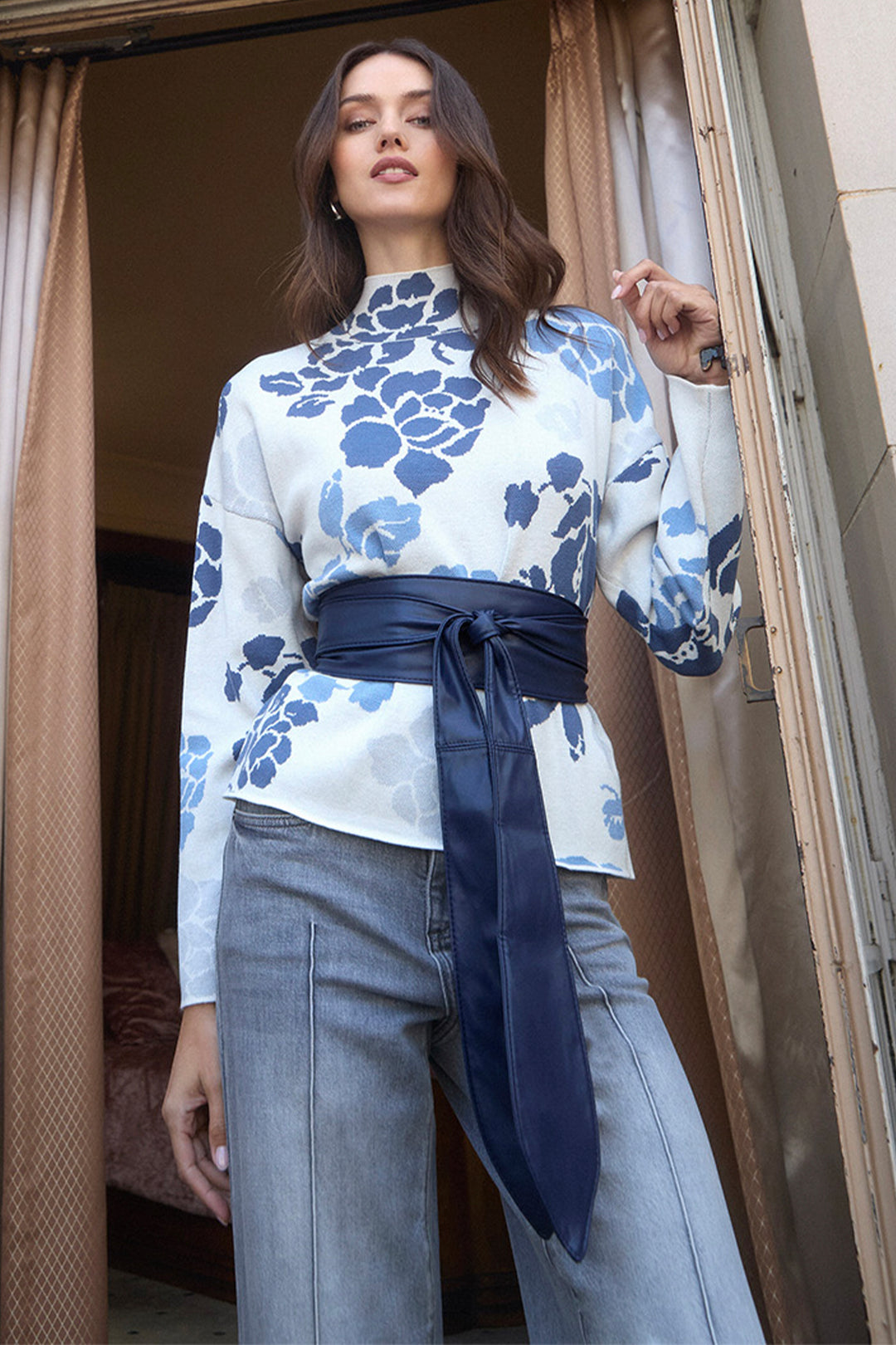 A woman models a long-sleeve mock-neck blouse with a blue floral pattern on a white base, styled with a wide navy satin waist tie and light-wash jeans, standing in a doorway.