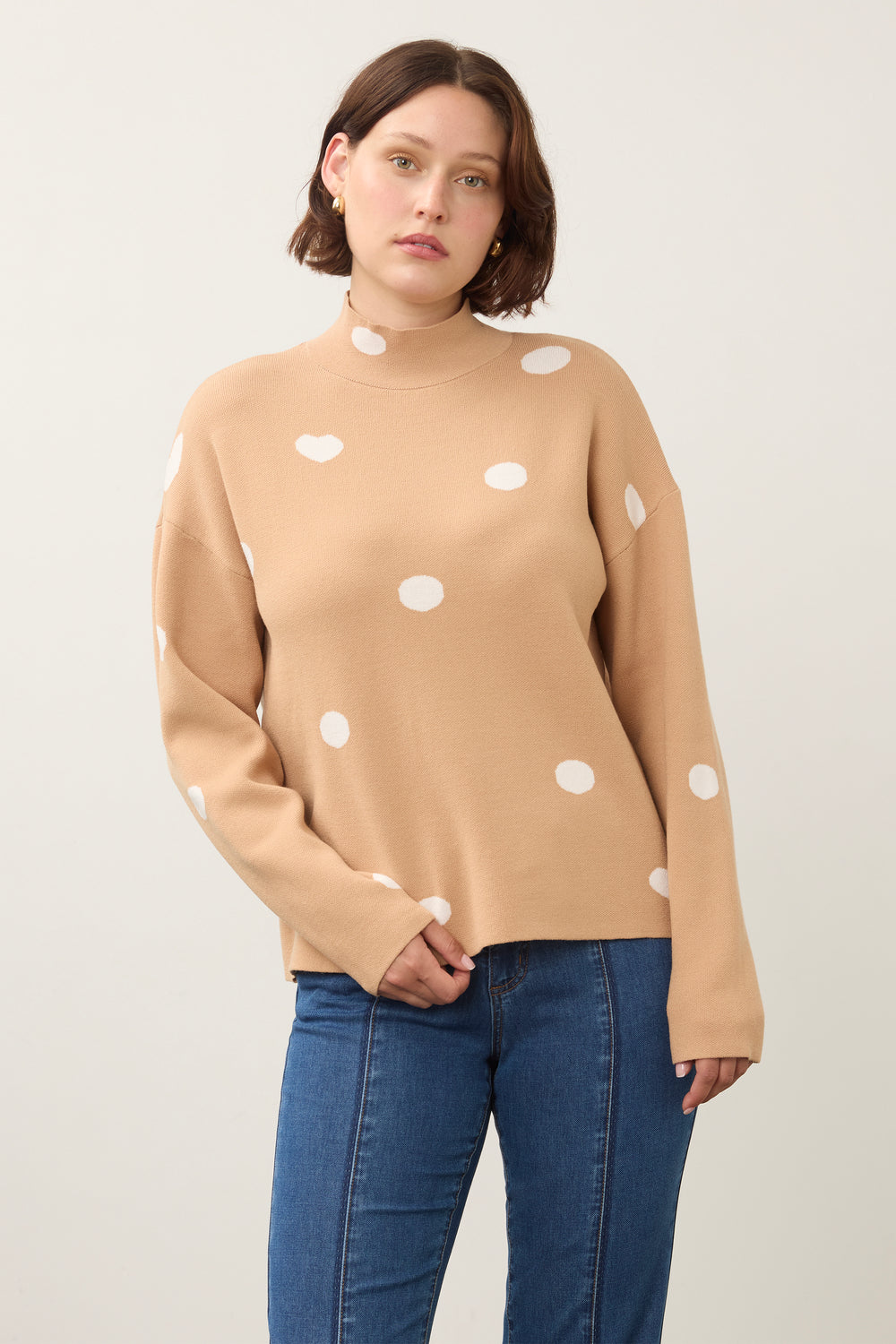 A woman wearing a camel-colored mock-neck knit sweater with white polka dots, paired with blue jeans, photographed against a plain light gray studio background.