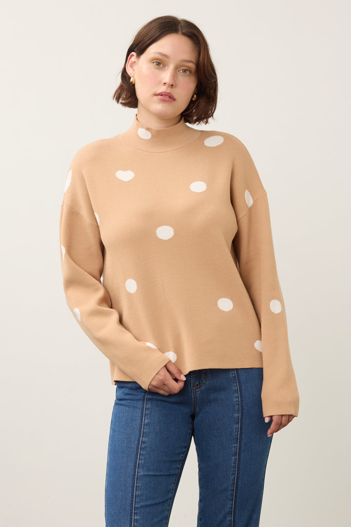 A woman wearing a camel-colored mock-neck knit sweater with white polka dots, paired with blue jeans, photographed against a plain light gray studio background.