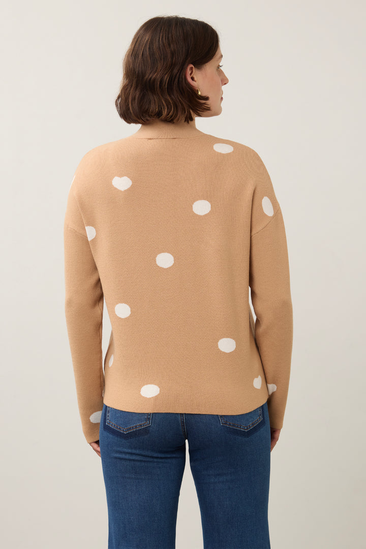 A woman viewed from the back wearing a camel-colored knit sweater with white polka dots and blue jeans, set against a plain light gray studio background.