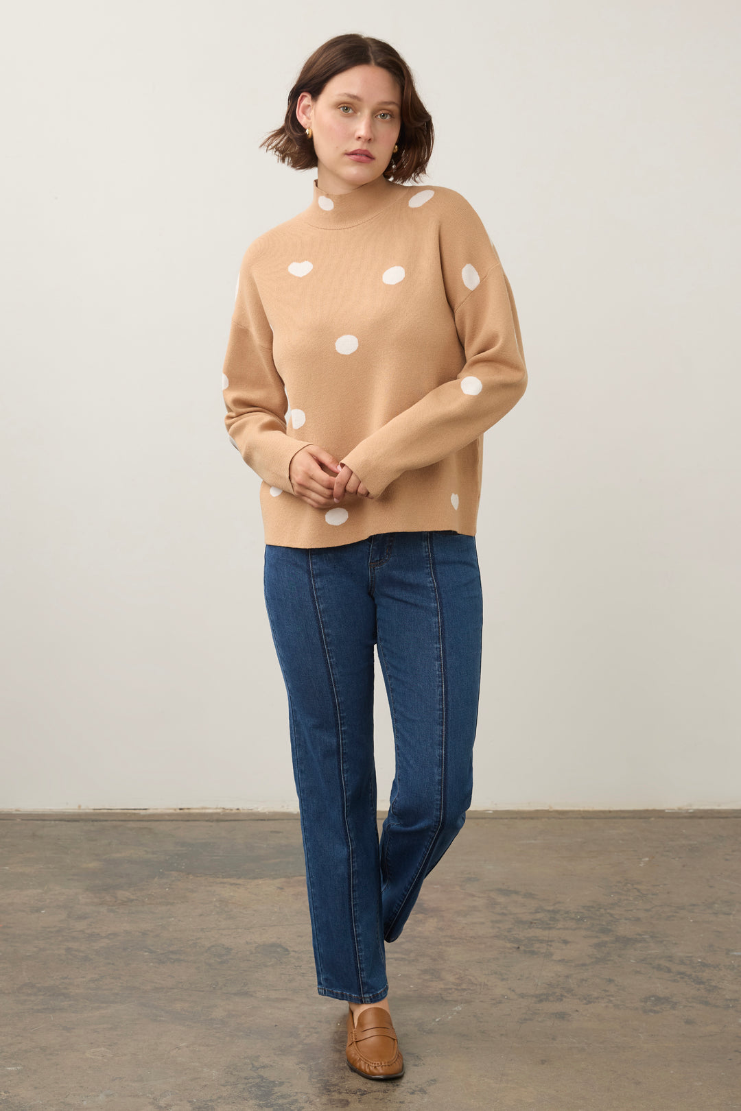 A woman modeling a beige mock-neck knit sweater with white polka dots, paired with dark blue jeans and brown leather loafers, standing against a plain off-white wall.