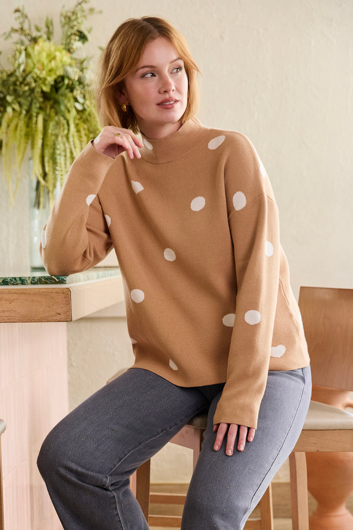 A woman wearing a camel-colored knit sweater with white polka dots, paired with gray jeans, sitting on a wooden stool in a brightly lit indoor setting.