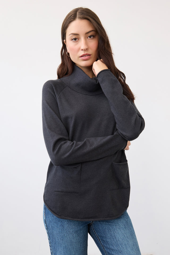 A woman modeling a dark charcoal gray long-sleeve turtleneck sweater with front patch pockets, paired with blue jeans, against a plain white studio background.