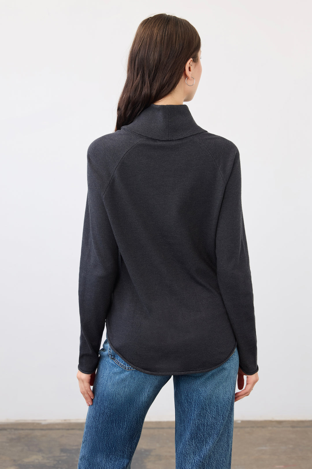 A woman viewed from the back wearing a dark charcoal gray mock neck long-sleeve top and blue jeans, standing against a plain light gray wall.