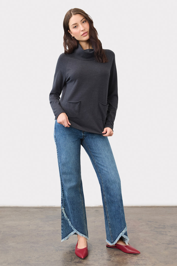 A woman modeling a dark charcoal gray mock neck knit sweater with front pockets, paired with wide-leg blue jeans featuring frayed hems and red pointed flats, set against a plain studio background.