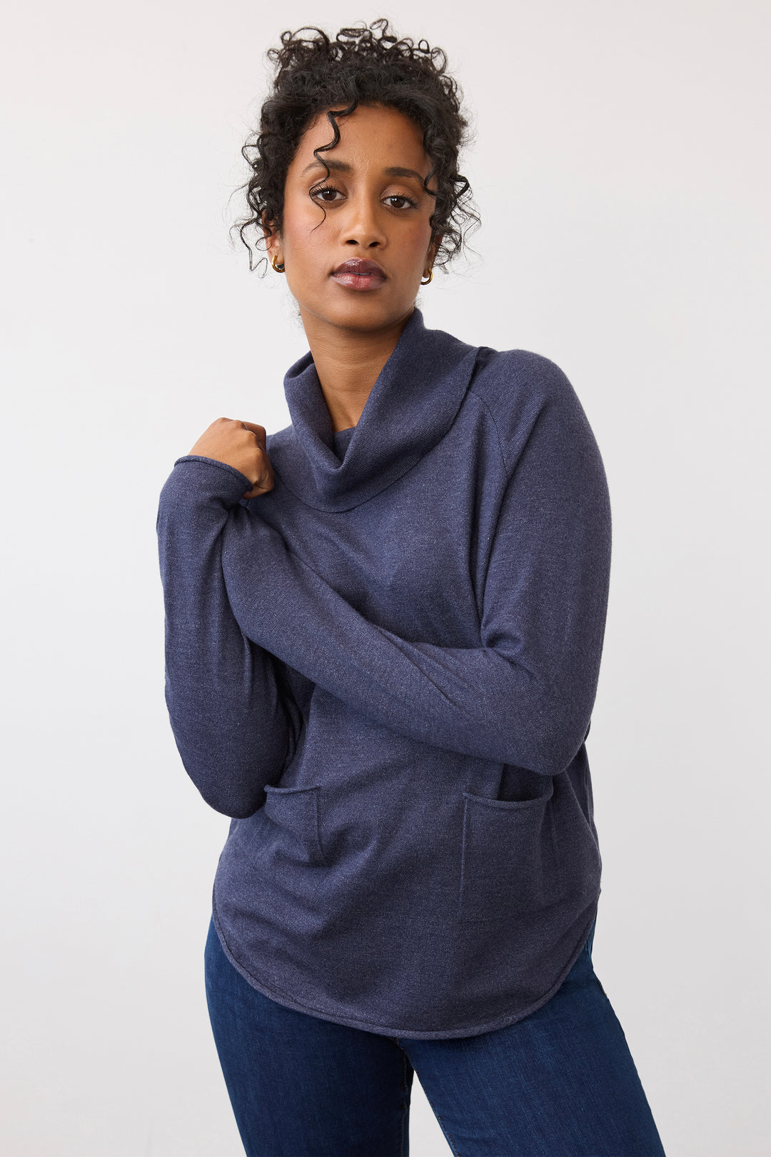A woman wearing a dark slate blue cowl neck long-sleeve top with front pockets, paired with dark blue jeans, posing against a light gray studio background.