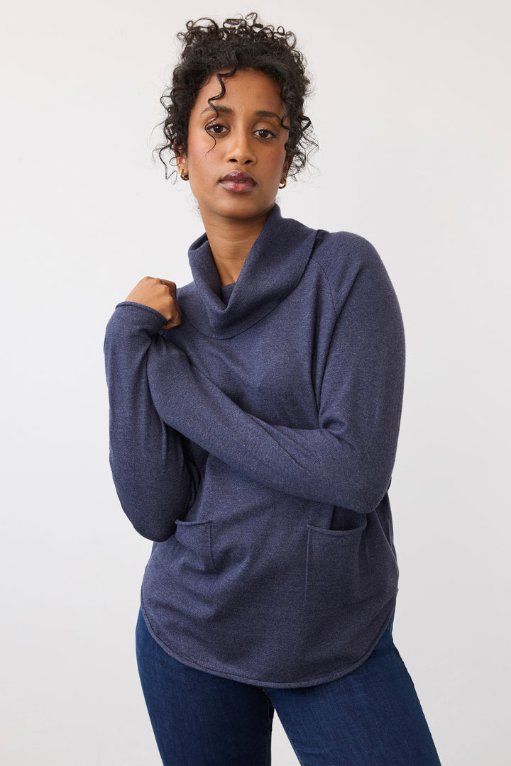 A woman wearing a dark slate blue cowl neck long-sleeve top with front pockets, paired with dark blue jeans, posing against a light gray studio background.
