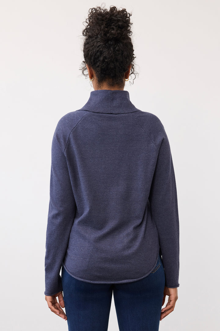 A woman viewed from the back wearing a heathered slate blue mock neck long-sleeve top and dark blue jeans against a plain white background.