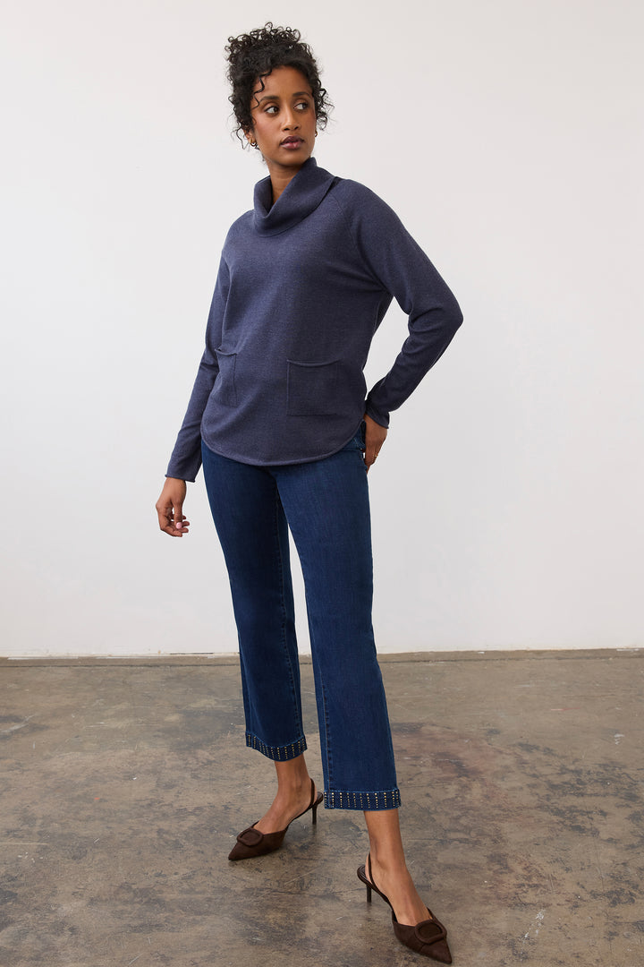 A woman modeling a navy blue long-sleeve cowl neck sweater with front patch pockets, paired with dark blue cropped jeans and brown pointed-toe slingback heels, standing against a plain off-white wall.