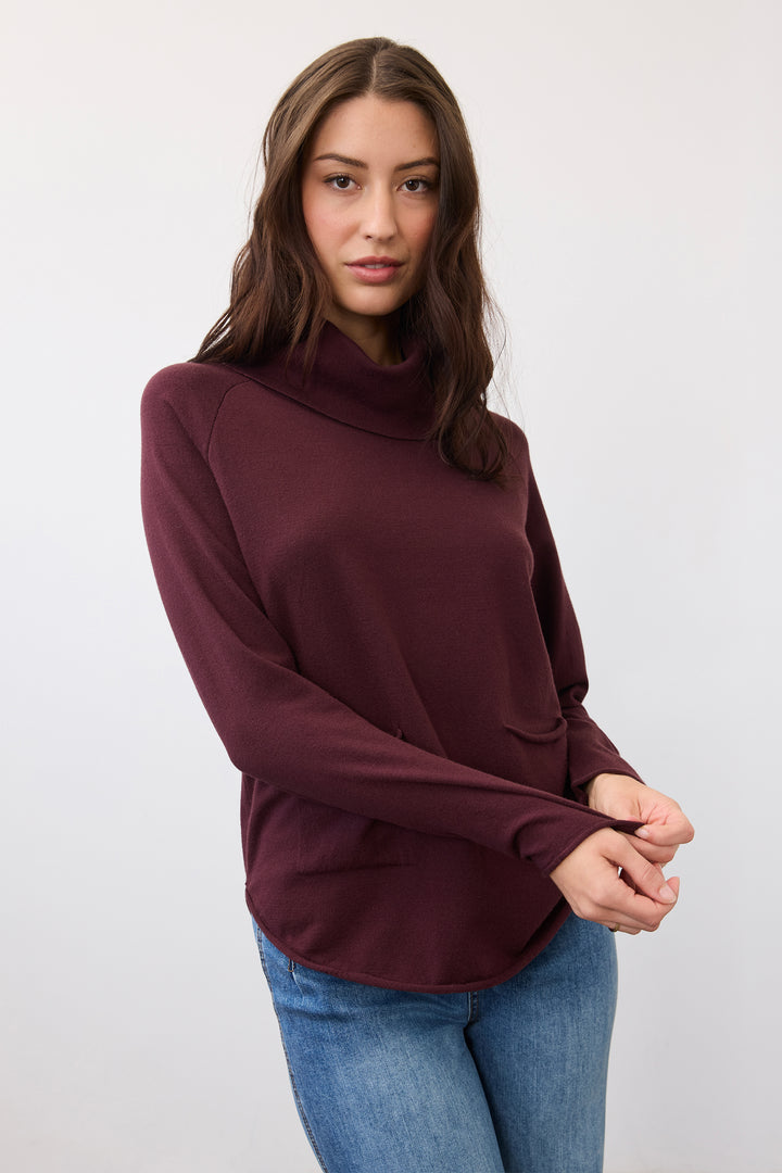 A woman wearing a long-sleeve, high-neck turtleneck top in deep burgundy with blue jeans, standing against a plain light gray studio background.