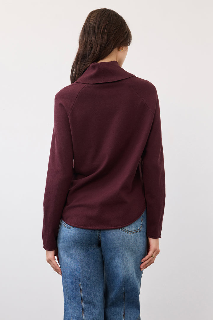 A woman viewed from the back wearing a dark burgundy long-sleeve mock neck top and blue jeans, standing against a plain light gray background.