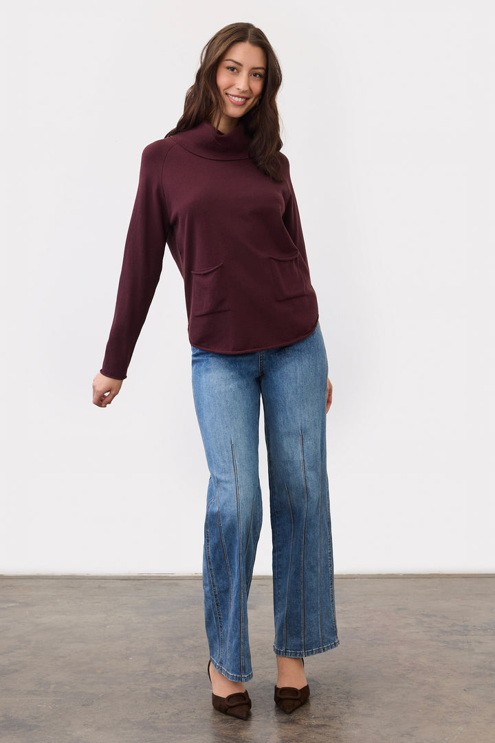 A woman modeling a long-sleeve, high-neck, deep burgundy knit top with front patch pockets, paired with blue jeans and brown shoes, standing against a plain white studio wall.