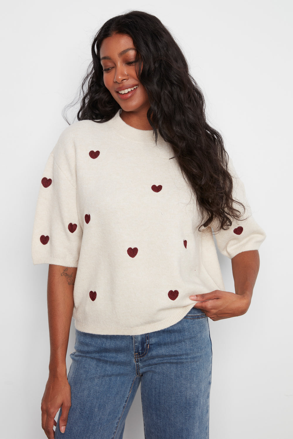 A woman wearing a cream-colored knit sweater with small dark red heart appliqués and blue jeans, standing against a plain white studio background.
