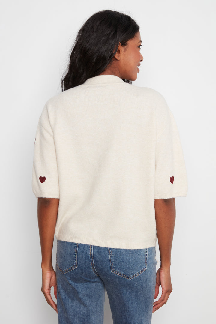 A woman viewed from the back wearing a cream-colored knit sweater with small red heart details on the sleeves, paired with blue jeans, against a plain white background.