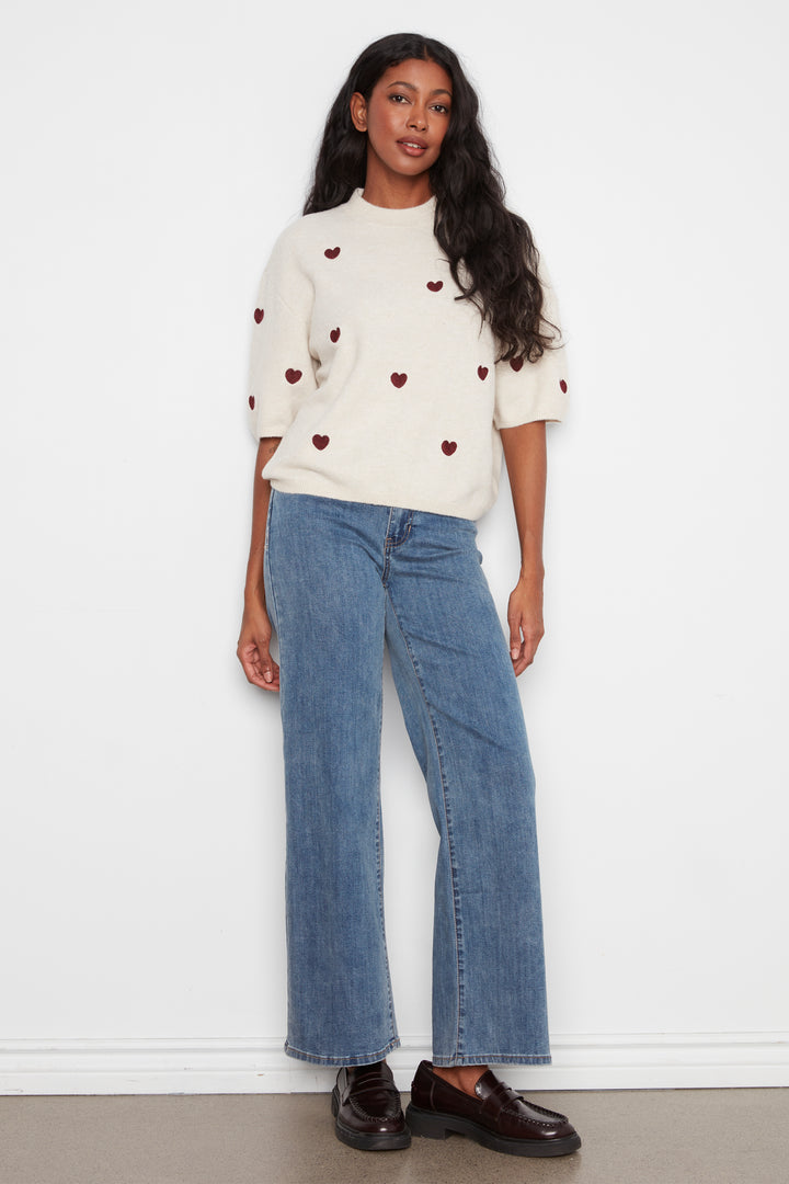 A woman modeling a cream-colored short-sleeve knit top with small red heart details, paired with wide-leg blue jeans and dark brown leather loafers, standing against a plain white wall.