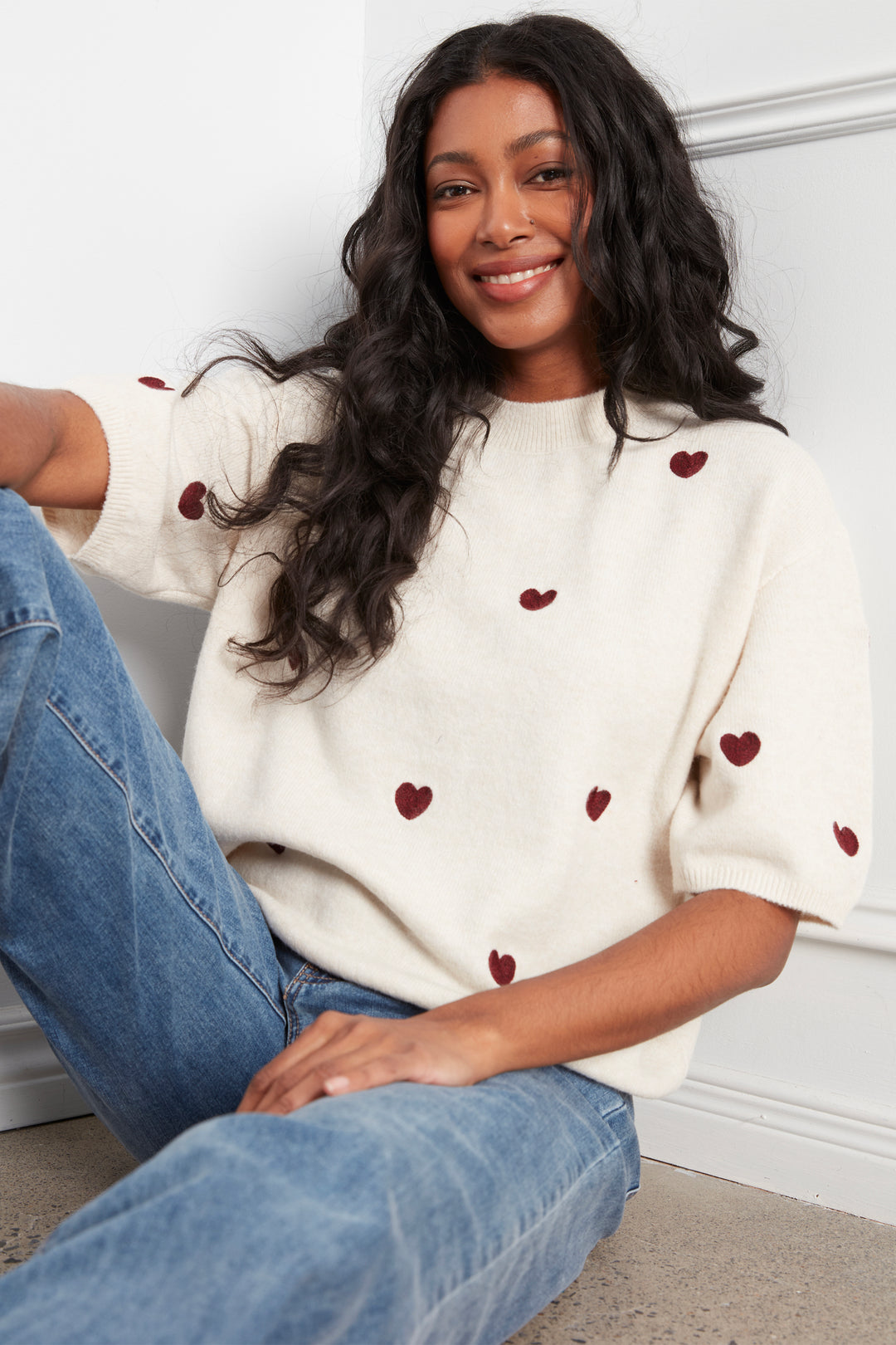 A woman wearing a cream-colored knit sweater with small red heart details, paired with blue jeans, sitting against a white paneled wall.