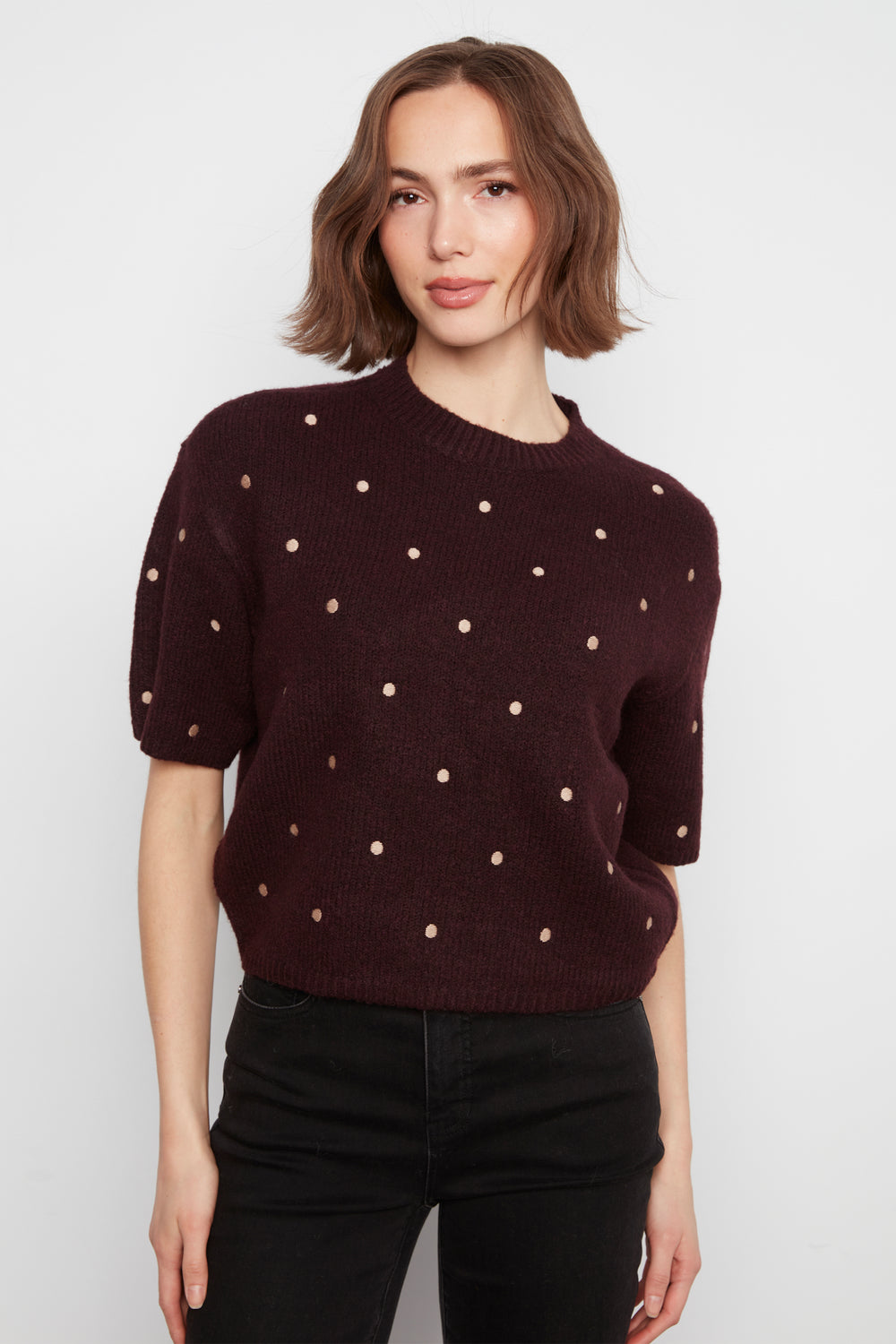 A woman wearing a short-sleeve crewneck sweater in deep burgundy with small metallic gold polka dots, paired with black pants, against a plain light gray background.