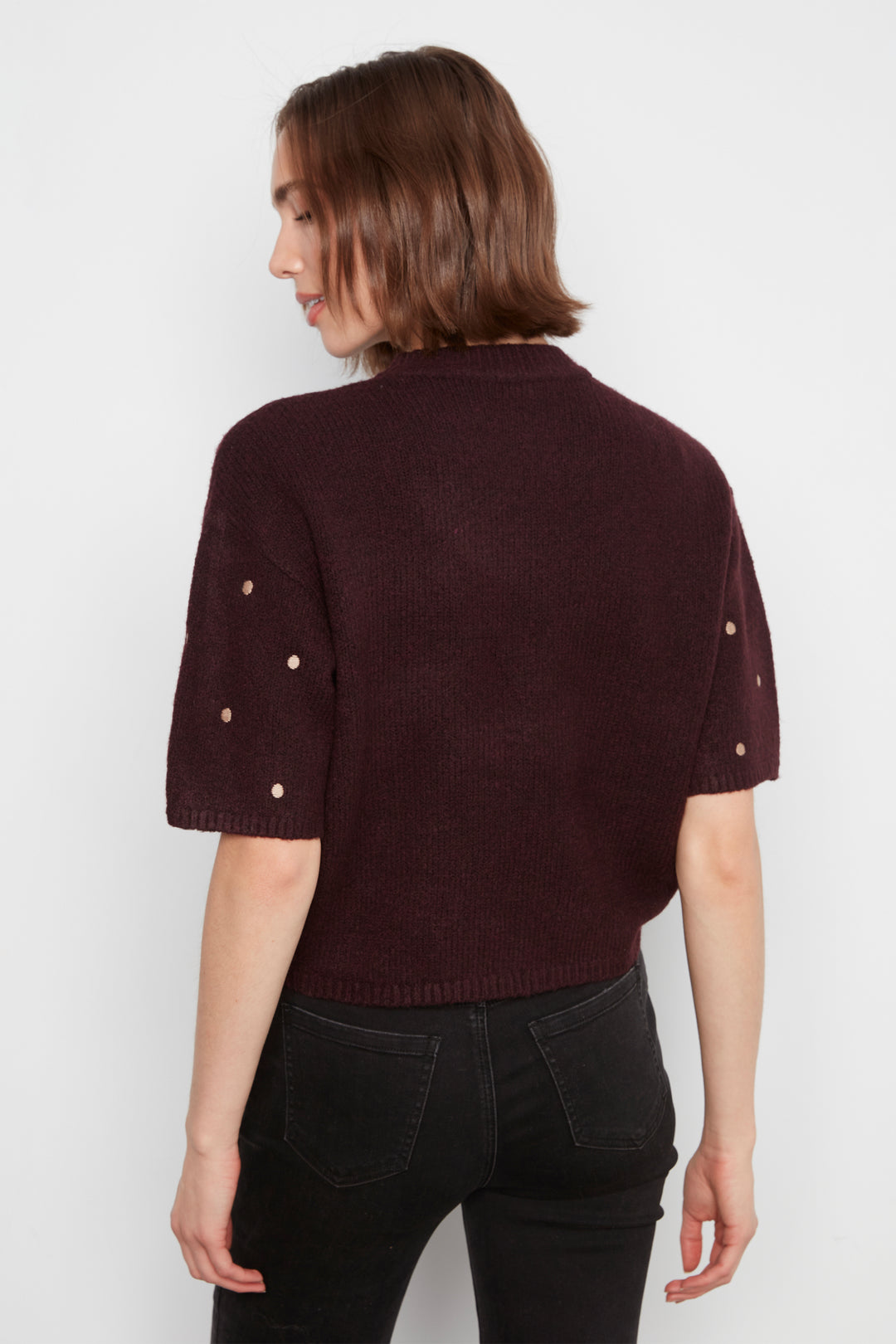 A woman shown from the back wearing a short-sleeve knit sweater in deep burgundy with gold-tone decorative buttons on the sleeves, paired with black jeans, against a plain light gray background.