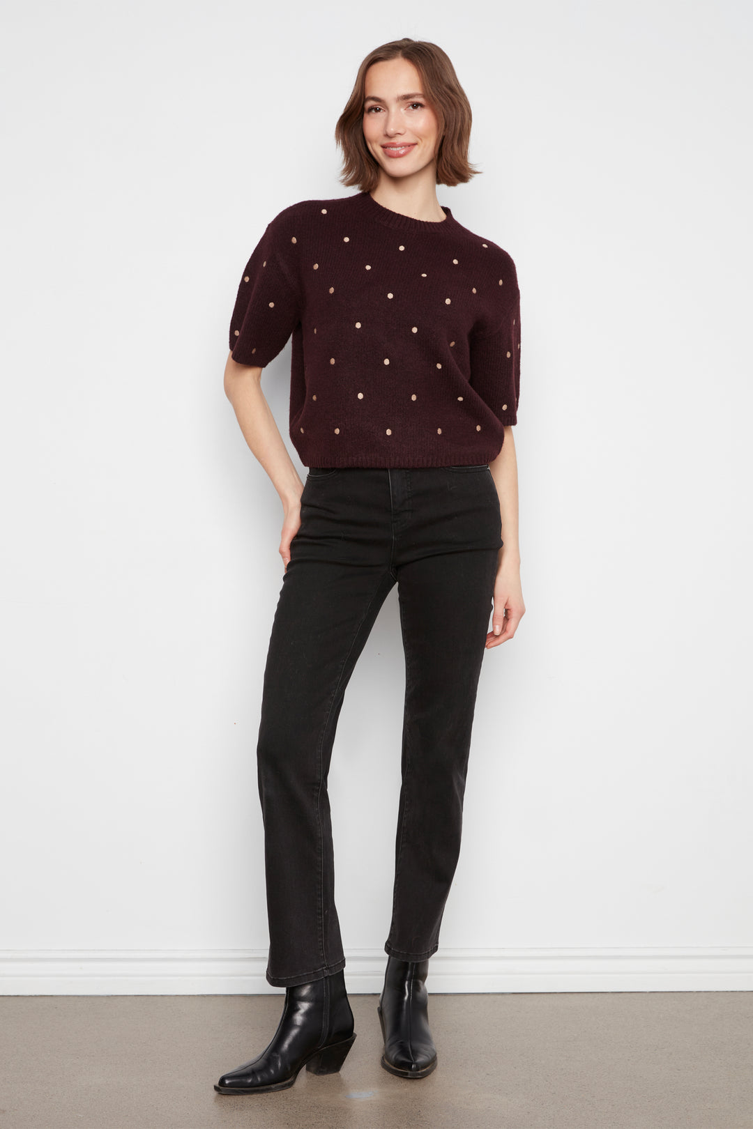 A woman modeling a dark burgundy short-sleeve knit sweater with small white embroidered dots, paired with black jeans and black ankle boots, standing against a white wall.