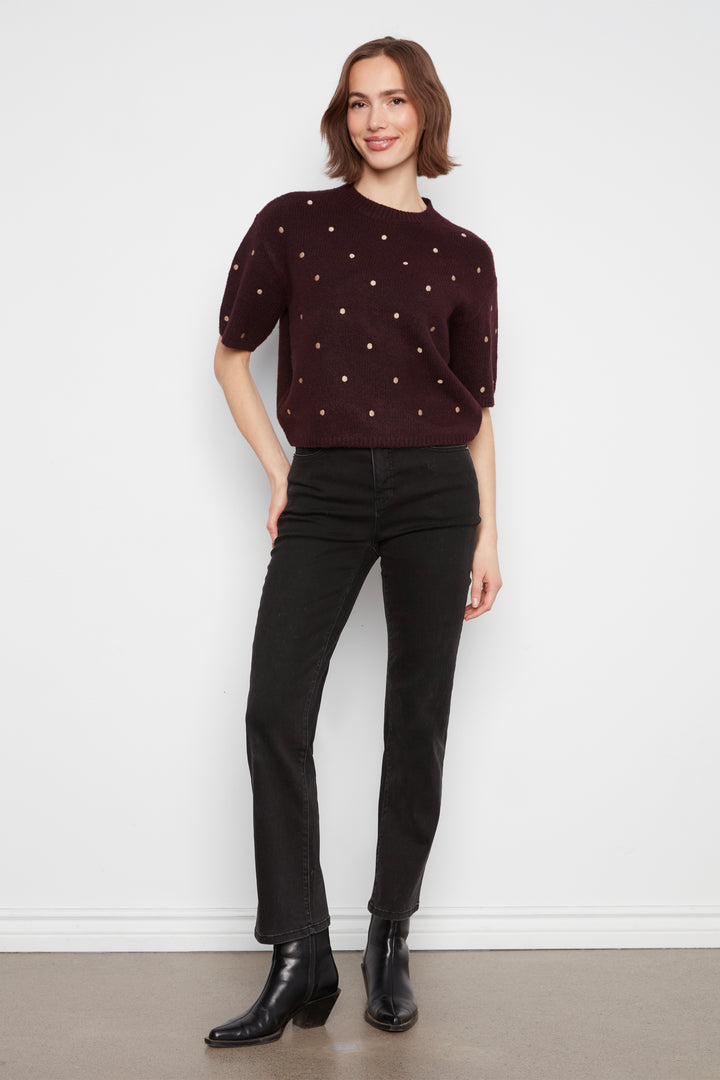 A woman modeling a dark burgundy short-sleeve knit sweater with small white embroidered dots, paired with black jeans and black ankle boots, standing against a white wall.