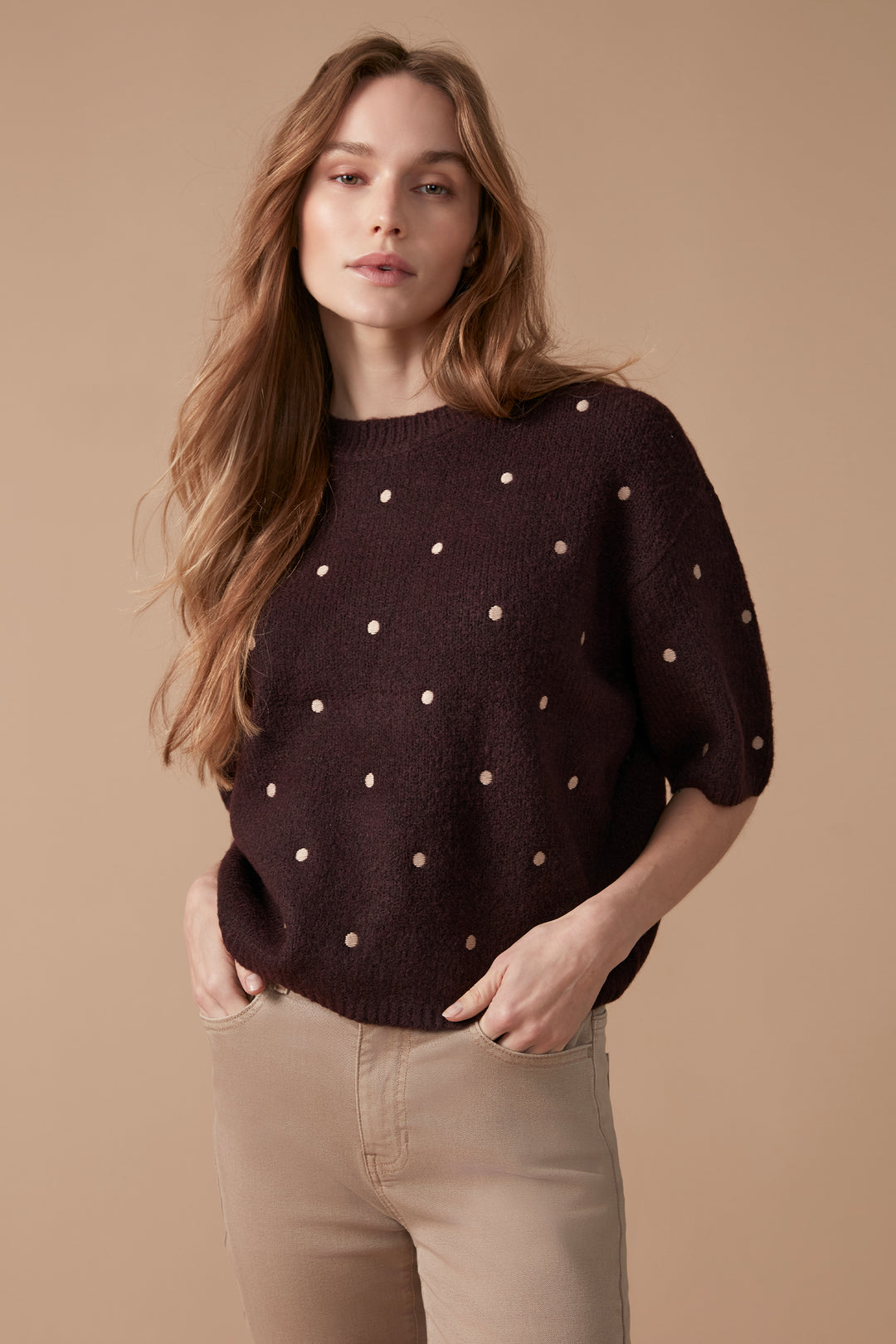 A woman modeling a dark brown knit sweater with small beige polka dots, paired with beige pants, against a solid beige studio background.