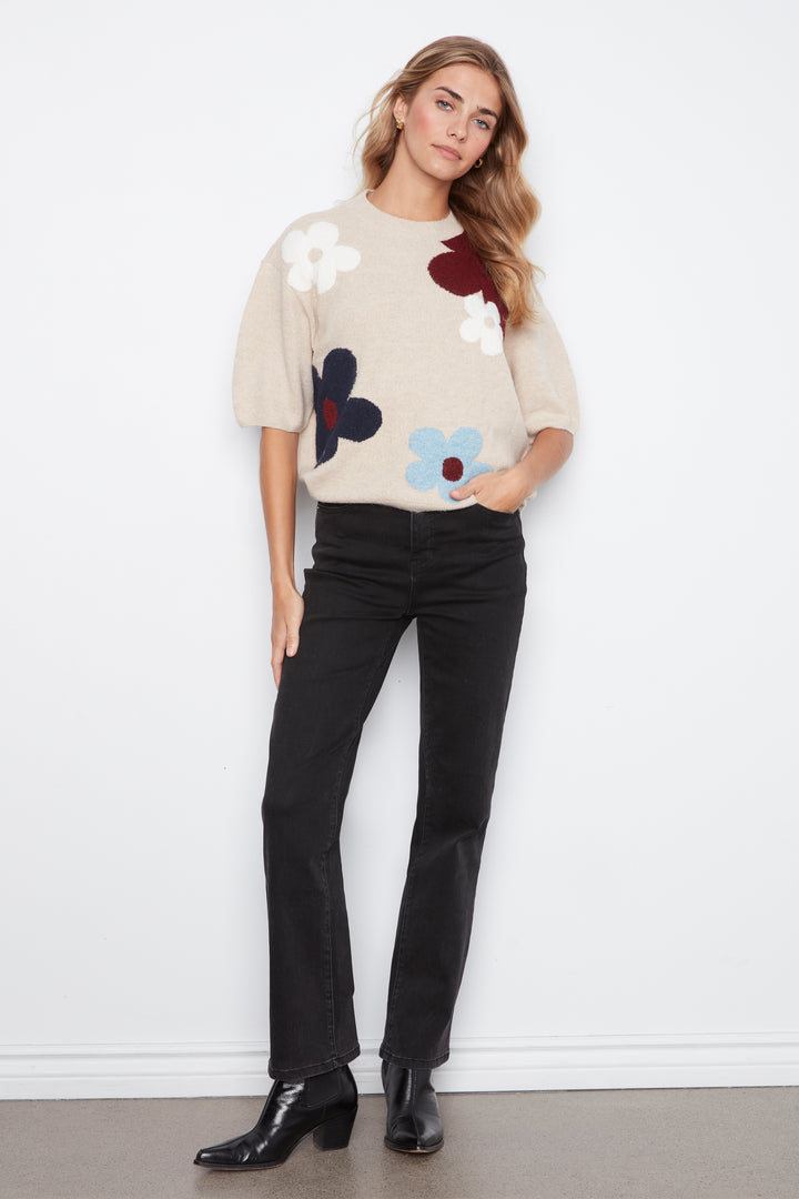 A woman modeling a beige short-sleeve knit sweater with large multicolor flower appliqués in white, dark blue, burgundy, and light blue, paired with black jeans and black ankle boots against a white wall.