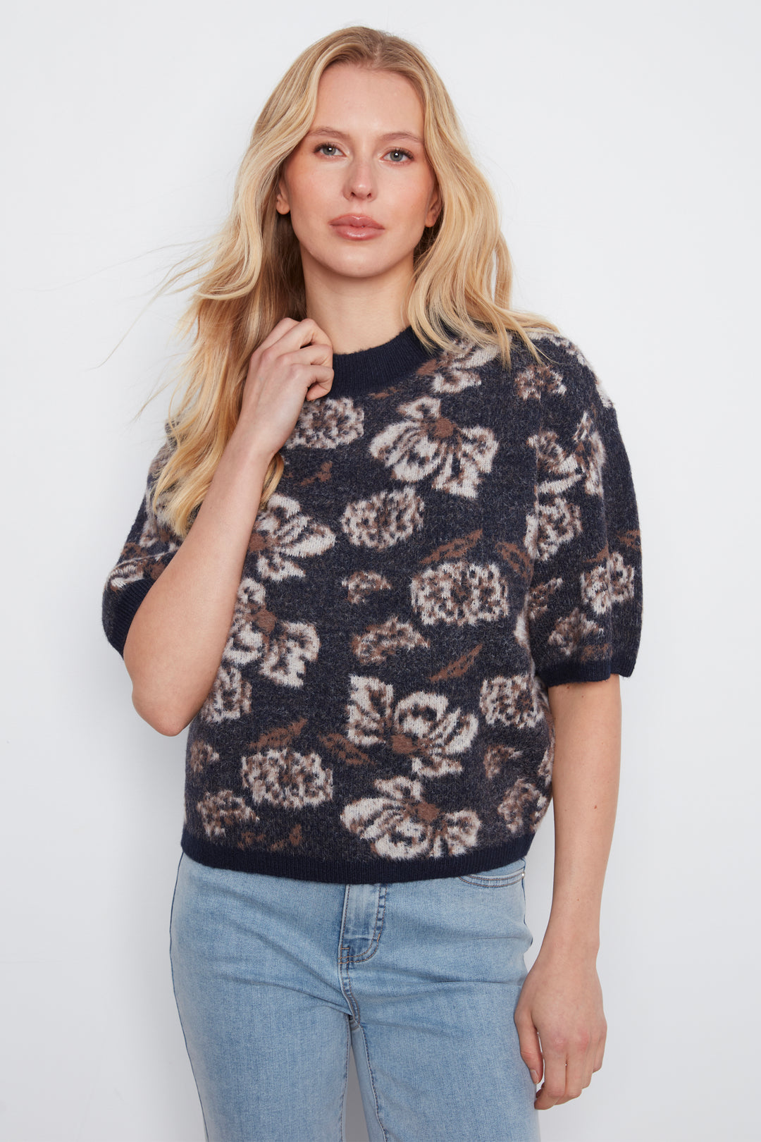 A blonde woman wearing a short-sleeve knit sweater with a dark navy base and a beige and brown floral pattern, paired with light blue jeans, against a plain white background.