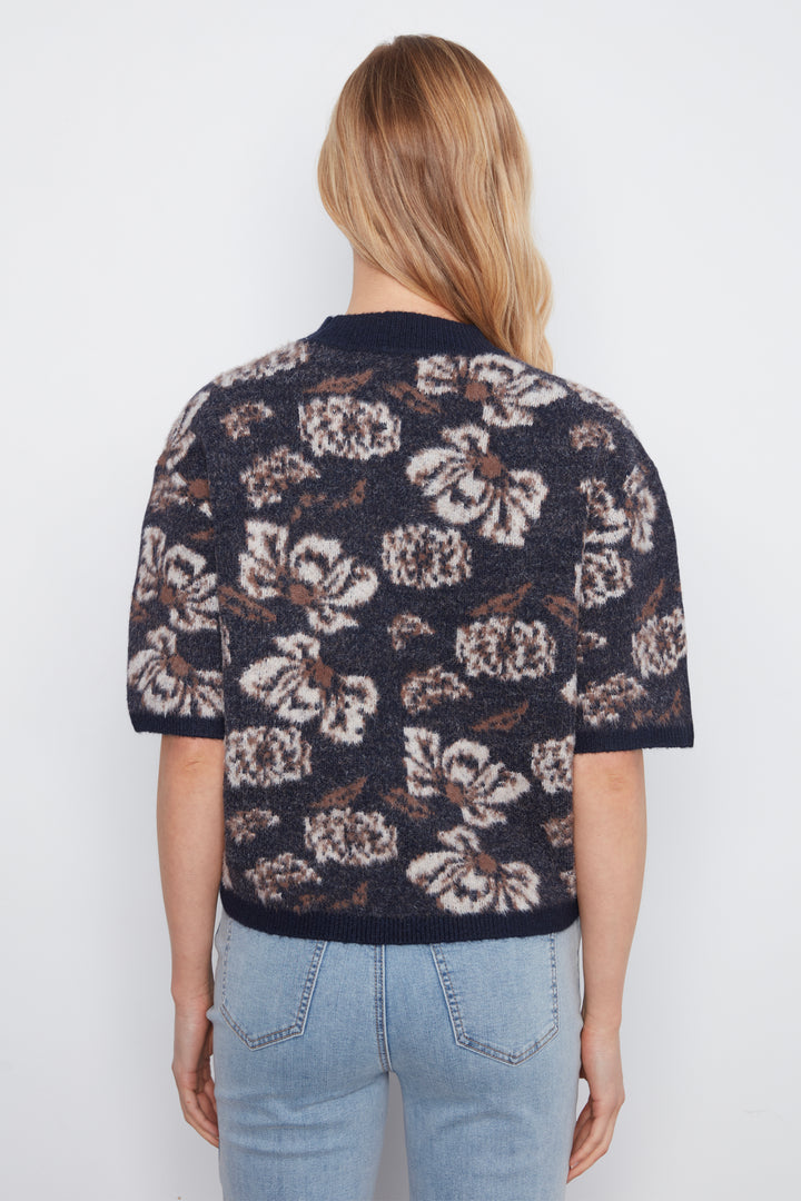 A woman viewed from behind wearing a short-sleeve knit top with a dark navy base and a large cream and brown floral pattern, paired with light blue jeans against a plain white background.