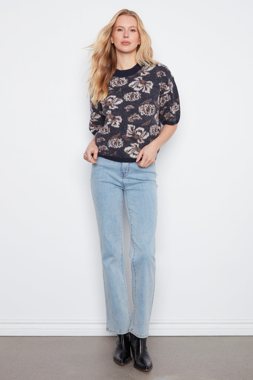 A woman stands against a white wall wearing a dark floral knit sweater with light blue straight-leg jeans and black ankle boots.