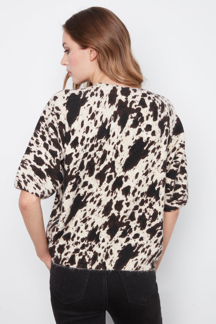 A woman shown from the back wearing a short-sleeve top with a black and off-white cow print pattern, paired with black jeans, against a plain light gray background.