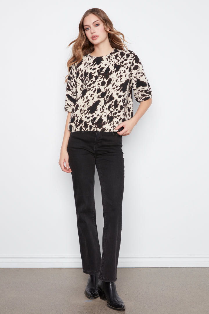 A woman modeling a beige and black abstract patterned knit top with three-quarter sleeves, paired with black jeans and black boots, standing against a white wall.
